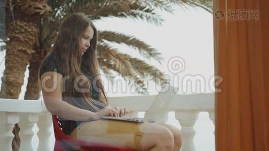 漂亮的女学生或在笔记本电脑上工作的自由职业者，坐在阳台上看棕榈和大海，速度很快视频