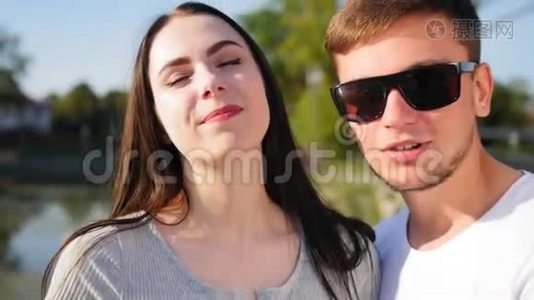 一对年轻幸福的夫妇看着镜头的肖像。 在池塘附近的绿色夏季公园里的浪漫情侣。 快乐视频