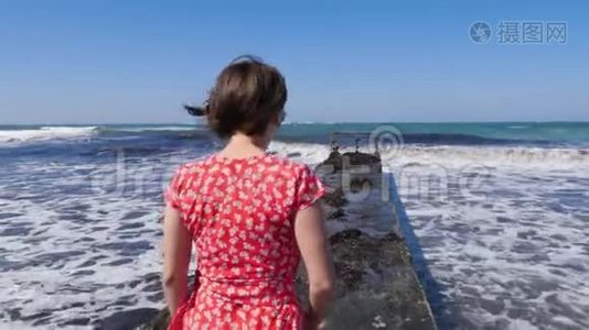 年轻迷人的白种人女人穿着红色的裙子走在码头上。 赤脚的双腿朝着码头上的大海走去.. 慢节奏视频