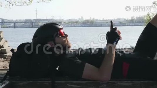 骑自行车的人穿着黑色运动服，坐在长凳上，手里拿着手机，太阳在太阳镜里反射。 带着头盔的线人用他的视频