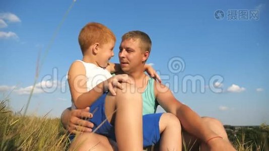 微笑的红发男孩和他的父亲坐在草地上的绿草，一起在户外玩。 快乐爸爸视频