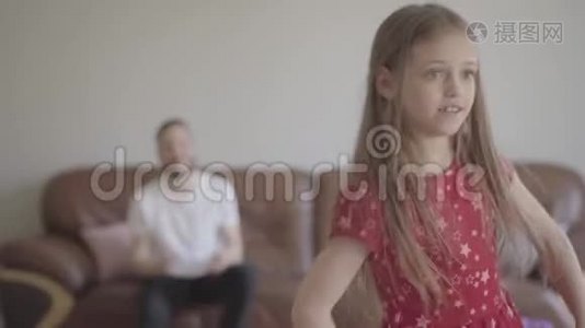 可爱的小滑稽女孩穿着红色连衣裙在蓬松的地毯上的前景中跳舞的肖像。 坐在那儿的父亲视频