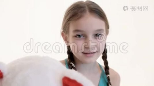 一个可爱的白色女孩的肖像，她扎着辫子，抱着一只大泰迪熊，坐在地板上，背景是白色的。视频