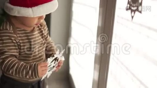 戴着圣诞帽的小宝宝在室内对着窗户玩圣诞装饰。 新年快乐，圣诞快乐视频