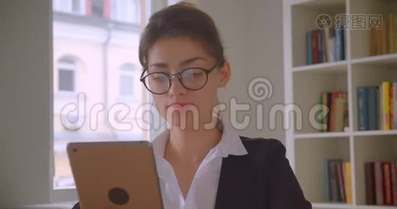 一位年轻漂亮的白种人女商人戴着眼镜在办公室室内的平板电脑上浏览特写镜头视频
