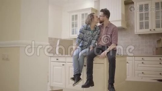 年轻的接吻夫妇坐在新公寓的厨房桌子上。 一家人搬到一个新家。 带着一个视频