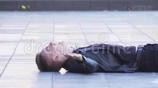 迷人的年轻金发男性穿着黑色夹克躺在白色瓷砖地板上。视频