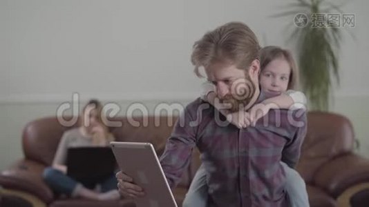 这位留胡子的人在平板电脑上工作，参与了他的工作。 悲伤的小女孩坐在父亲的背上，试图得到视频