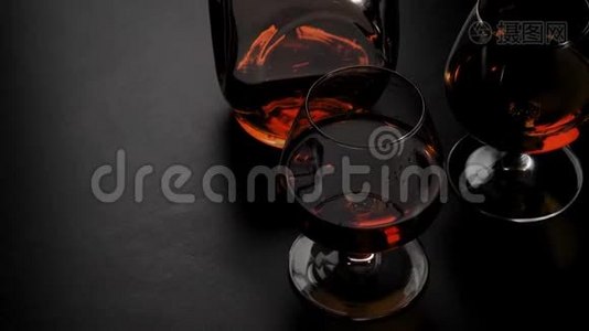 豪华白兰地。 带有金色干邑的圆形瓶子和两个玻璃杯在黑色背景下的黑色桌子上旋转。 玻璃眩光。视频