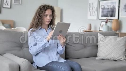 坐在沙发上的卷发女士在平板电脑上网上购物视频