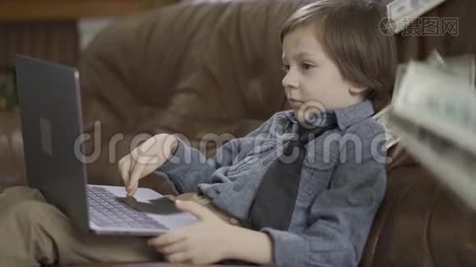 那个穿着牛仔裤夹克的小男孩坐在皮革沙发上玩笔记本电脑。 一美元落在沙发上，孩子视频