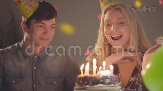 可爱的微笑金发女孩戴着生日帽吹灭蛋糕上的蜡烛，年轻人坐在附近。 女人有视频