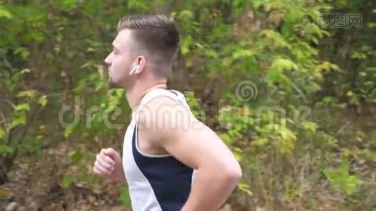初秋，一个运动的年轻人沿着森林附近的小径奔跑。 带无线耳机的帅哥慢跑视频