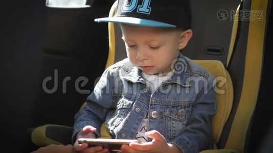 可爱的蹒跚学步的小男孩坐在汽车座椅上，从智能手机上观看视频。 小孩在车里玩智能手机视频