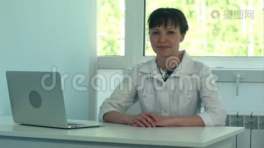 女医生带着笔记本电脑在办公桌上工作，对着相机微笑视频
