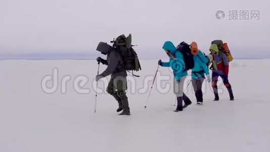 一群活跃的男人和女人带着背包和滑雪杆在强烈的冬季风中穿过冰冻的河流。视频