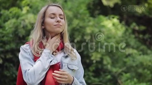 生病的年轻女子戴着红领巾感觉不好，喉咙痛，站在公园里。 痛苦的吞咽视频