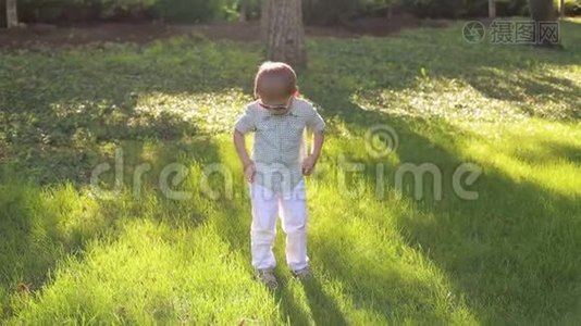 一个穿着白色裤子、衬衫和太阳镜的小男孩站在绿草上视频