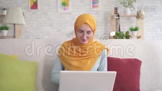 积极的年轻穆斯林妇女，手提电脑坐在客厅的沙发上，看着相机微笑视频