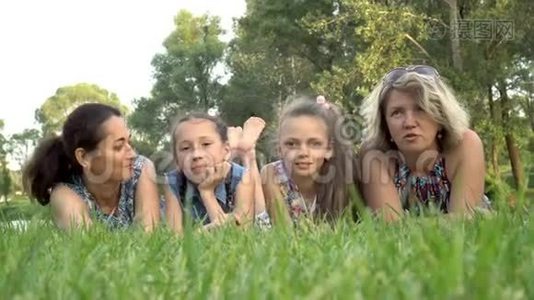 日落时分，两位母亲和两个小女儿躺在公园的绿草上，玩耍和亲吻。 肖像。 关闭视频