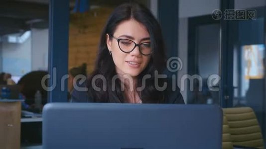 商务女性感到快乐，微笑着，看着办公室的镜头。 年轻女性在办公桌上使用笔记本电脑视频