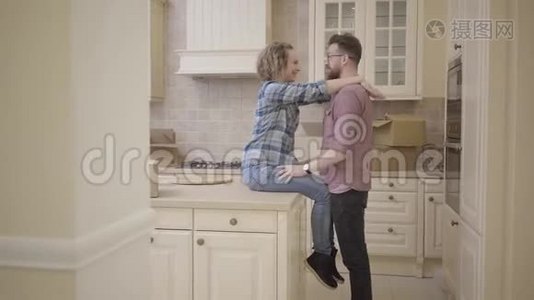 大胡子男人站在漂亮女人旁边，坐在半空的大厨房里的桌子上。 女人拥抱她的丈夫，他们接吻视频