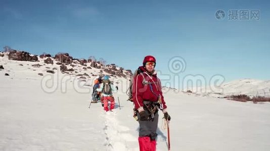 穿着红色温暖的西装站在雪地里，手里拿着冰镐，望着远处。 在他身后视频