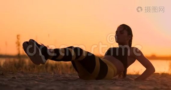 年轻美丽的女子运动员在海滩上练习，在日落时为腹肌的肌肉做运动。 扭扭捏捏视频