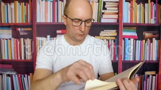 体贴的白人在图书馆内部的书架背景下读书。 戴着眼镜和白色t恤的男人坐在视频