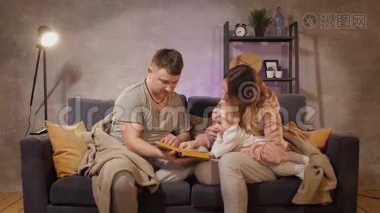 一个年轻的家庭坐在舒适的房间里的沙发上。 妈妈，爸爸和宝宝在看书.. 家庭幸福视频