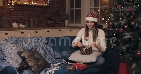 快乐的年轻美丽的女人，戴着桑塔帽，坐在靠近圣诞树的沙发上，在舒适的圣诞节背景下。视频