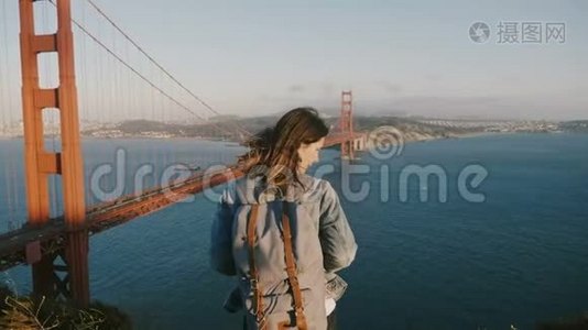 镜头放大年轻幸福的女子背后背包，头发在风中欣赏日落风景在金门大桥。视频