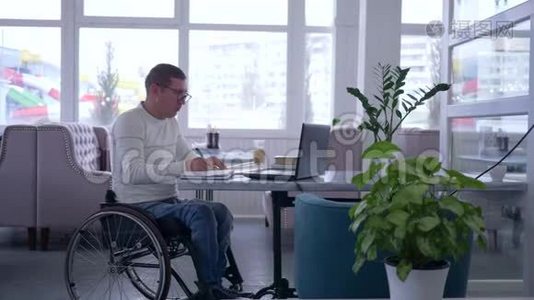 电子商务，成功的成熟男性，轮椅上的残疾人戴眼镜，使用笔记本电脑坐在咖啡馆的桌子上视频