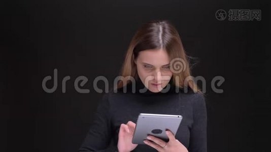 年轻的白种人长发金发的金发女孩聚精会神地看着平板电脑，在黑色背景下微笑着进入相机。视频