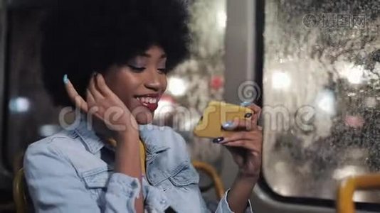 微笑着年轻的非洲裔美国妇女看有趣的视频智能手机骑在公共交通。 夜间视频