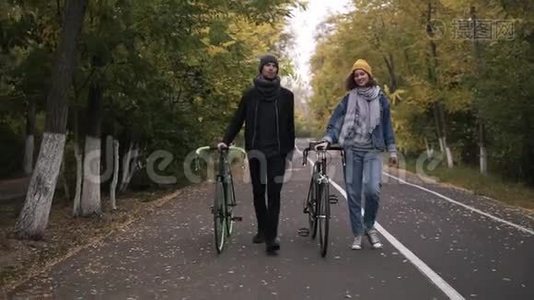 两位亲密的朋友在绿黄高树之间的城市公园里骑着自行车散步，聊得很开心视频