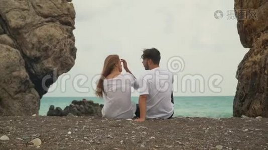 年轻的一对夫妇坐在两块石头之间的美丽海滩上，交谈着视频