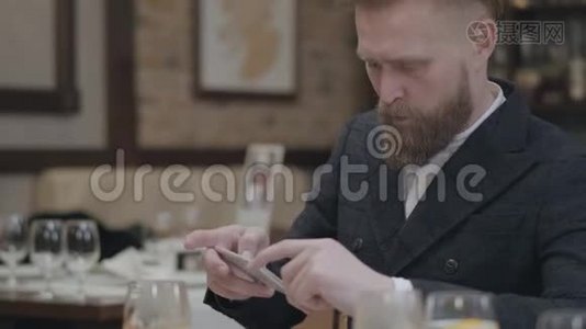 自信的金发大胡子男，穿着时髦的夹克，拿着手机坐在一个现代化的房间里拍照视频