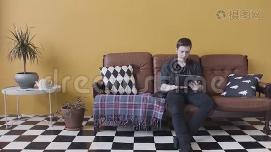 英俊的休闲年轻人在家里使用平板电脑，坐在客厅的沙发上，浏览互联网。 股票视频