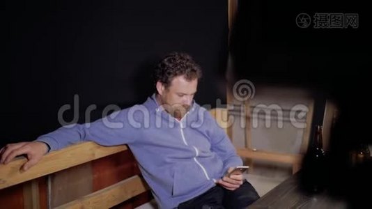 一个留着胡子和胡子的人正坐在桌旁用手机写短信，面前的桌上放着两瓶视频