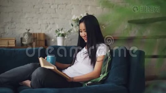 美丽的亚洲女人正在看书，在舒适的阁楼式公寓里坐在沙发上，享受着有趣的故事和欢笑视频