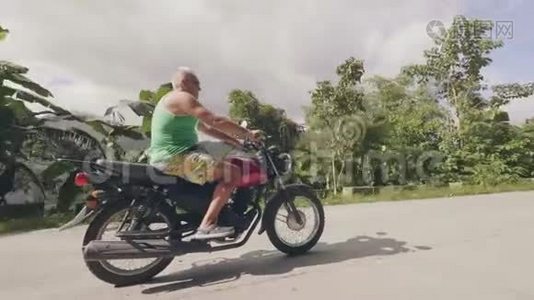成熟男子骑摩托车骑摩托车在乡村道路上的热带棕榈树景观。 高级官员旅行视频