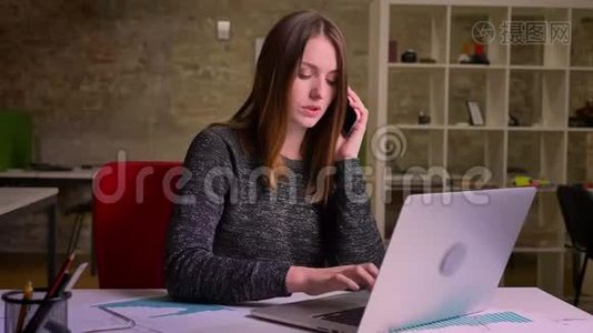 年轻的红头发的女商人在笔记本电脑前工作，用手机说话，做笔记视频