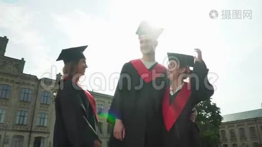 三个穿着黑色和红色壁炉的快乐学生在大学大楼前庆祝他们的毕业。 两个女孩视频