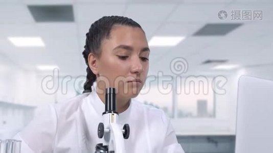 一位女性研究人员在显微镜下观察生物样本，并在坐在白色的笔记本电脑前将数据写入电脑视频