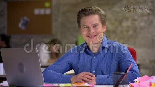 中年金发商人在笔记本电脑前的肖像，在办公室里对着镜头微笑和眨眼。视频