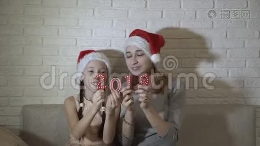 两个戴着圣诞`帽子的小女孩拿着2019年的红色数字，坐在沙发上，跳舞，微笑着视频