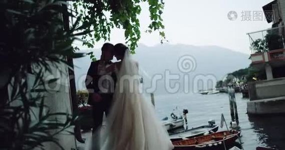 漂亮的新娘穿着长长的裙子，新郎在迷人的地方亲吻和拥抱，旁边有湖和船视频