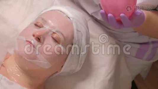 美容师在一家美容院用白色刷子将凝胶通过面膜敷在女性脸上。 女人的皮肤视频