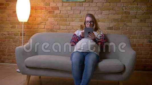 年轻怀孕女性坐在舒适的室内公寓沙发上浏览平板电脑的特写镜头视频